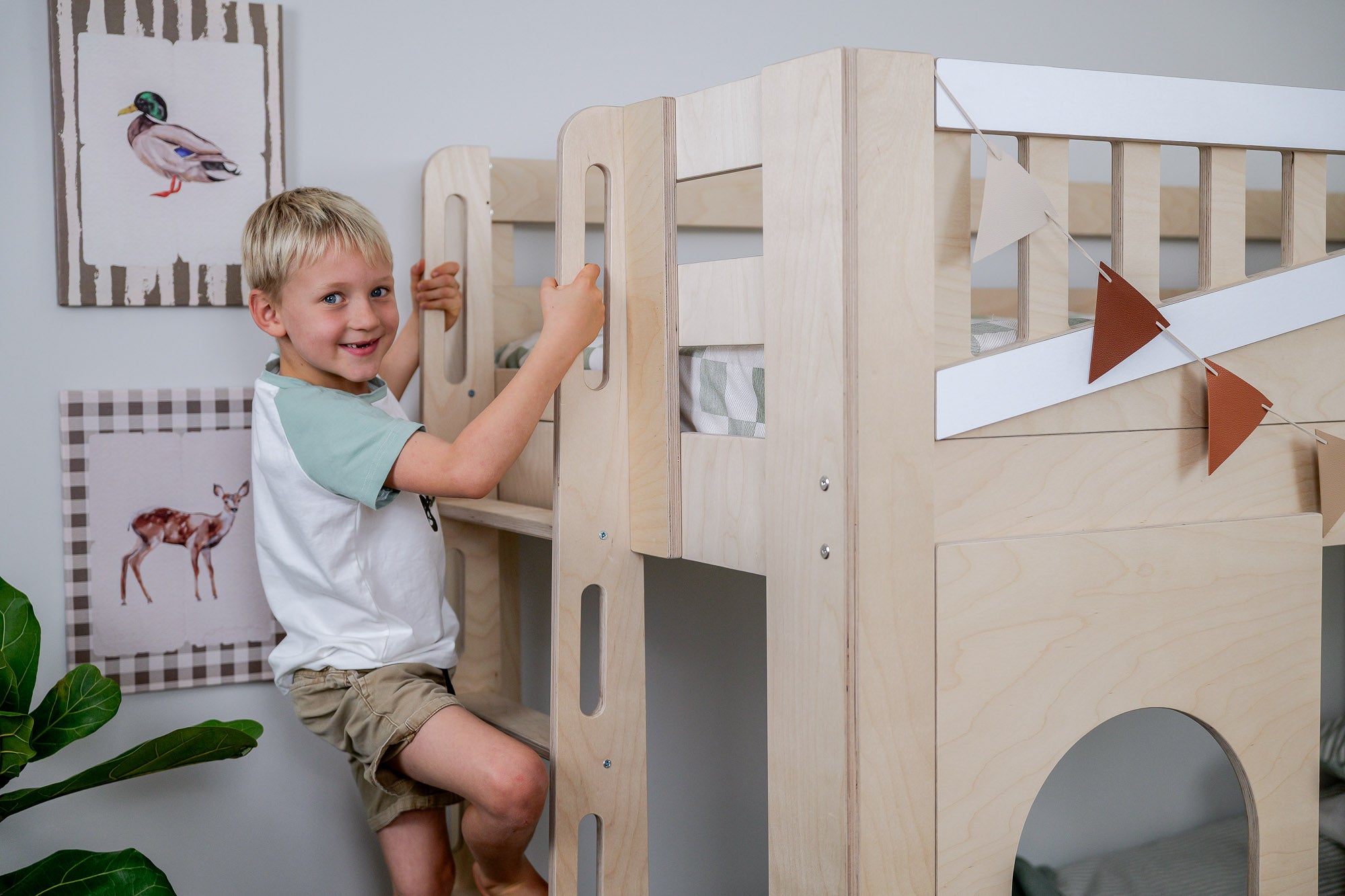 Child playing on a wooden bunk bed in a room with framed pictures on the wall.