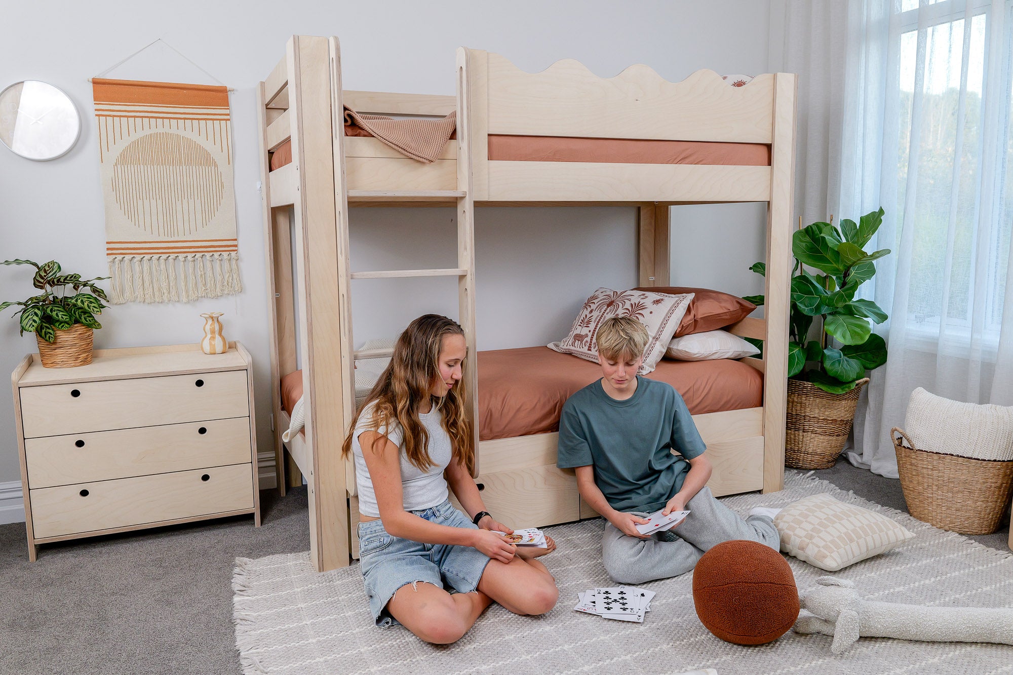 Two children sitting on the floor next to a set of bunk beds in a room with plants and furniture.